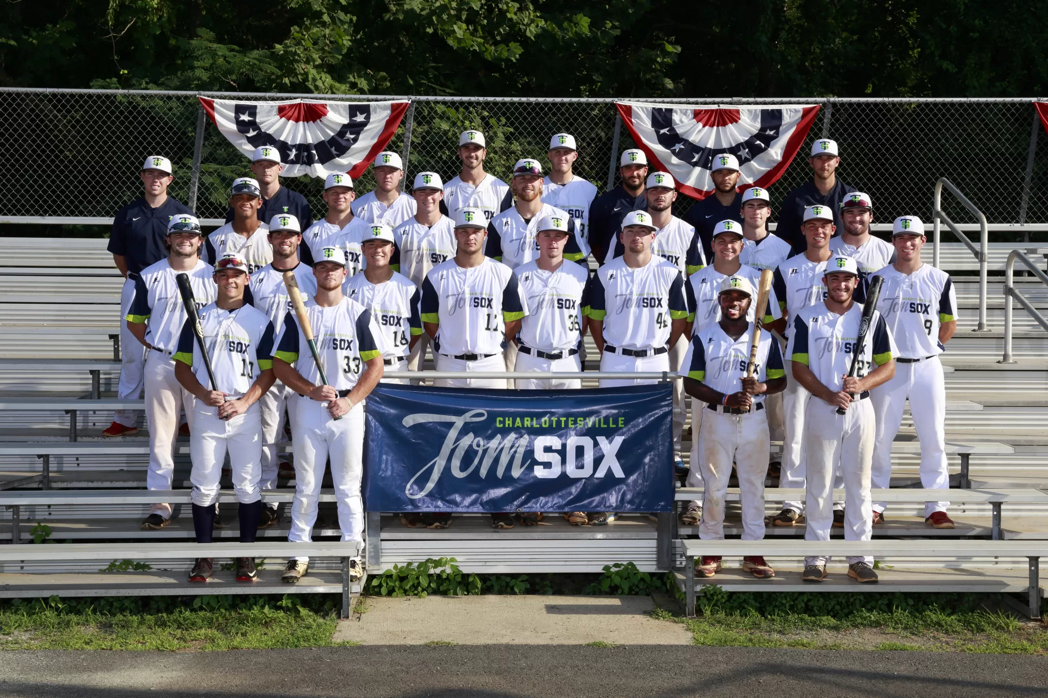 Photo of the Tom Sox team lined up on the bleachers. The middle features the Tom Sox logo and there is bunting in the back. The players are wearing white jerseys.